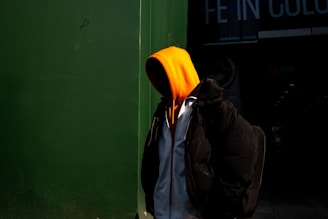 A person wearing an orange hooded sweatshirt stands partially in shadow against a dark green wall. The person appears to be wearing a black jacket over the sweatshirt, with a backpack visible on their back. Bright contrast creates a dramatic effect with the vivid orange hood standing out.