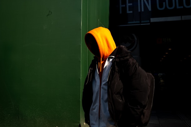 A person wearing an orange hooded sweatshirt stands partially in shadow against a dark green wall. The person appears to be wearing a black jacket over the sweatshirt, with a backpack visible on their back. Bright contrast creates a dramatic effect with the vivid orange hood standing out.