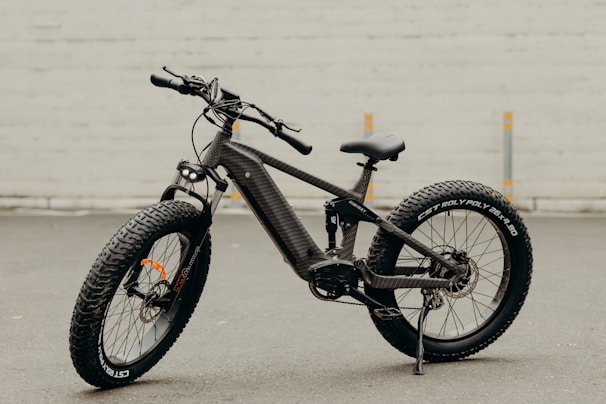 A black, modern fat-tire electric bike is positioned against a smooth, pale background. The bike features thick, rugged tires suitable for off-road terrain and a sleek carbon fiber frame. The battery and motor are integrated into the frame, highlighting its electric capability.