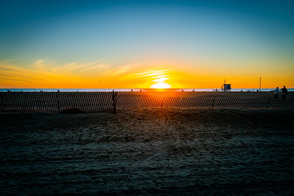 o sol está se pondo sobre a praia com uma cerca em primeiro plano