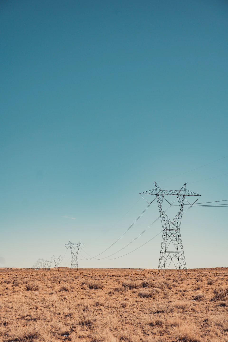 The image depicts a vast, open field with two tall, gray power lines running across it from left to right. The sky above is a clear blue, providing a striking contrast against the earthy tones of the field and power lines. In the distance, there are several other power lines that stretch out into the horizon, creating an intricate network of electricity across the landscape.
