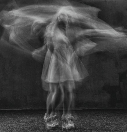 A blurred, ghost-like figure of a woman in motion, captured in black and white. The woman's movement creates a layered effect, suggesting swift dance or motion amidst a dark background.