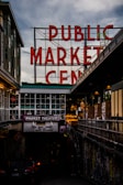 A large neon sign with red letters spelling out 'PUBLIC MARKET CENTER' stands prominently against a cloudy sky. Below is a market theater entrance with a marquee advertising live improv comedy. The surrounding area is urban, with nearby buildings and an alley filled with colorful graffiti.
