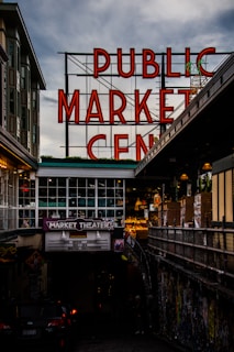 A large neon sign with red letters spelling out 'PUBLIC MARKET CENTER' stands prominently against a cloudy sky. Below is a market theater entrance with a marquee advertising live improv comedy. The surrounding area is urban, with nearby buildings and an alley filled with colorful graffiti.
