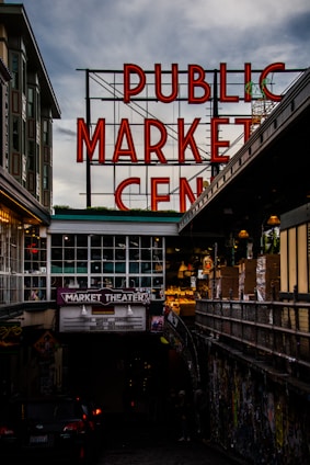 A large neon sign with red letters spelling out 'PUBLIC MARKET CENTER' stands prominently against a cloudy sky. Below is a market theater entrance with a marquee advertising live improv comedy. The surrounding area is urban, with nearby buildings and an alley filled with colorful graffiti.