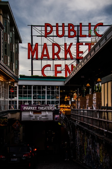 A large neon sign with red letters spelling out 'PUBLIC MARKET CENTER' stands prominently against a cloudy sky. Below is a market theater entrance with a marquee advertising live improv comedy. The surrounding area is urban, with nearby buildings and an alley filled with colorful graffiti.