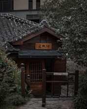 A traditional Japanese-style wooden house with a tiled roof is nestled among lush greenery. The structure features a wooden sign with Japanese characters and a prominent entrance gate. The surrounding foliage includes a variety of plants and bushes, contributing to a peaceful garden setting.