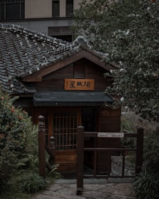 A traditional Japanese-style wooden house with a tiled roof is nestled among lush greenery. The structure features a wooden sign with Japanese characters and a prominent entrance gate. The surrounding foliage includes a variety of plants and bushes, contributing to a peaceful garden setting.