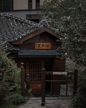 A traditional Japanese-style wooden house with a tiled roof is nestled among lush greenery. The structure features a wooden sign with Japanese characters and a prominent entrance gate. The surrounding foliage includes a variety of plants and bushes, contributing to a peaceful garden setting.