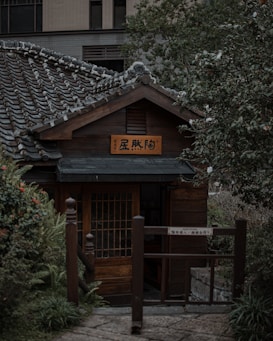 A traditional Japanese-style wooden house with a tiled roof is nestled among lush greenery. The structure features a wooden sign with Japanese characters and a prominent entrance gate. The surrounding foliage includes a variety of plants and bushes, contributing to a peaceful garden setting.