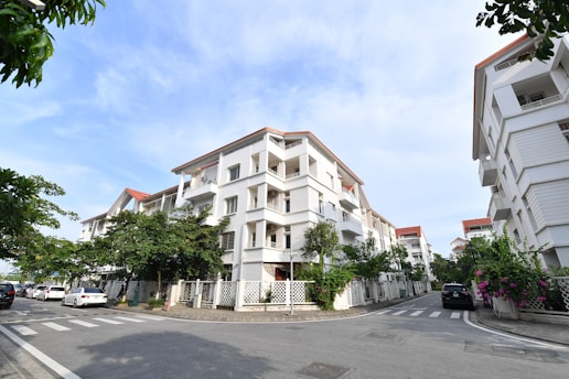 A residential area featuring modern white apartment buildings with red-tiled roofs and balconies. The buildings line a street with neatly paved roads and crosswalks. There are several parked cars by the roadside. Greenery and flowering plants are visible along the fences, adding a touch of nature to the urban setting. The sky is clear and blue, indicating a pleasant day.