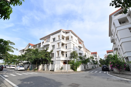 A residential area featuring modern white apartment buildings with red-tiled roofs and balconies. The buildings line a street with neatly paved roads and crosswalks. There are several parked cars by the roadside. Greenery and flowering plants are visible along the fences, adding a touch of nature to the urban setting. The sky is clear and blue, indicating a pleasant day.