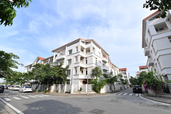 A residential area featuring modern white apartment buildings with red-tiled roofs and balconies. The buildings line a street with neatly paved roads and crosswalks. There are several parked cars by the roadside. Greenery and flowering plants are visible along the fences, adding a touch of nature to the urban setting. The sky is clear and blue, indicating a pleasant day.
