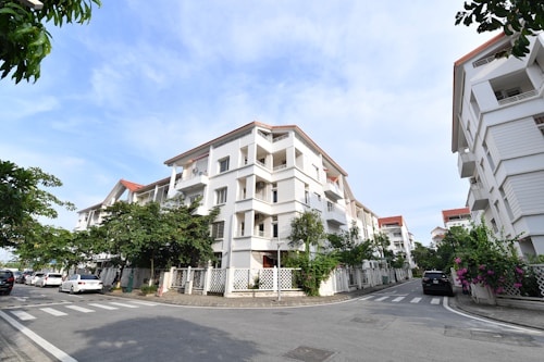 A residential area featuring modern white apartment buildings with red-tiled roofs and balconies. The buildings line a street with neatly paved roads and crosswalks. There are several parked cars by the roadside. Greenery and flowering plants are visible along the fences, adding a touch of nature to the urban setting. The sky is clear and blue, indicating a pleasant day.