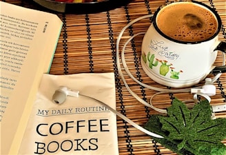 An arrangement on a wooden surface includes a book with visible text, a white mug filled with coffee, an illustrated design of cacti with the phrase 'Live your dream', a packet labeled 'My Daily Routine Coffee Books', a pair of white earphones, and a green leaf-shaped coaster.