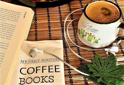 An arrangement on a wooden surface includes a book with visible text, a white mug filled with coffee, an illustrated design of cacti with the phrase 'Live your dream', a packet labeled 'My Daily Routine Coffee Books', a pair of white earphones, and a green leaf-shaped coaster.