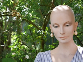 A mannequin head with a realistic face and large pearl earrings stands in front of lush green foliage under natural lighting. The mannequin wears a checkered garment, and small flowers can be seen among the leaves in the background.
