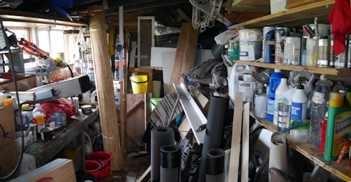 A cluttered workshop or garage space filled with various tools, paint cans, cleaning supplies, and miscellaneous items. Shelves are stacked with containers and bottles, while workbenches hold tools and other gear. The area is tightly packed with materials such as wood planks, rolls of fabric or carpet, and buckets.