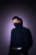 A person wears a dark turtleneck that covers most of their face, with only the eyes visible. They are standing against a softly lit purple background. The light creates a shadow on the wall, adding depth to the scene.