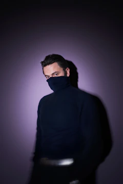 A person wears a dark turtleneck that covers most of their face, with only the eyes visible. They are standing against a softly lit purple background. The light creates a shadow on the wall, adding depth to the scene.