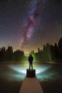 A person stands on a platform at the edge of a frozen lake, gazing up at the vibrant and expansive starry night sky. The Milky Way is prominently visible, stretching across the sky with rich details in purple, pink, and blue hues. Surrounding the scene are tall, dark trees, creating a silhouette against the softly lit horizon. A light source appears near the base of the platform, casting a glow on the icy surface.