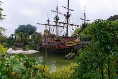 A large, ornate pirate ship with red and black coloring is docked near lush greenery and palm trees. The ship features multiple masts with unfurled sails and decorative golden accents, suggesting a historical, adventurous theme. Surrounding vegetation frames the scene, creating a contrast with the cloudy sky.