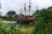 A large, ornate pirate ship with red and black coloring is docked near lush greenery and palm trees. The ship features multiple masts with unfurled sails and decorative golden accents, suggesting a historical, adventurous theme. Surrounding vegetation frames the scene, creating a contrast with the cloudy sky.
