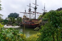 A large, ornate pirate ship with red and black coloring is docked near lush greenery and palm trees. The ship features multiple masts with unfurled sails and decorative golden accents, suggesting a historical, adventurous theme. Surrounding vegetation frames the scene, creating a contrast with the cloudy sky.
