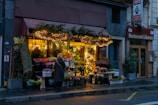 A cozy flower shop is adorned with string fairy lights. Outside, various colorful flowers and plants are on display. A person with a coat and scarf stands in front of the shop, adding a touch of warmth to the wet street scene. Next to the shop is a Lebanese restaurant with visible signage.