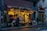 A cozy flower shop is adorned with string fairy lights. Outside, various colorful flowers and plants are on display. A person with a coat and scarf stands in front of the shop, adding a touch of warmth to the wet street scene. Next to the shop is a Lebanese restaurant with visible signage.