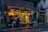 A cozy flower shop is adorned with string fairy lights. Outside, various colorful flowers and plants are on display. A person with a coat and scarf stands in front of the shop, adding a touch of warmth to the wet street scene. Next to the shop is a Lebanese restaurant with visible signage.