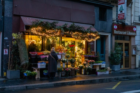 A cozy flower shop is adorned with string fairy lights. Outside, various colorful flowers and plants are on display. A person with a coat and scarf stands in front of the shop, adding a touch of warmth to the wet street scene. Next to the shop is a Lebanese restaurant with visible signage.
