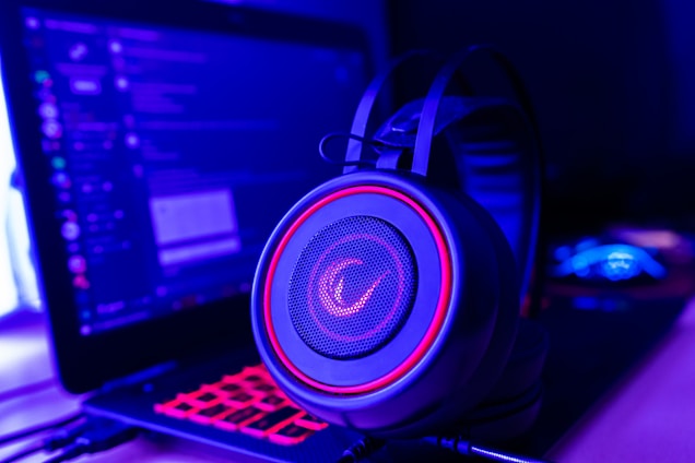 A pair of gaming headphones sits in front of a laptop displaying a chat application. The scene is bathed in a combination of blue and purple lighting, giving a high-tech and immersive atmosphere. The headphones have glowing red accents, enhancing the futuristic vibe.