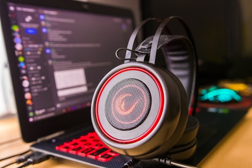 A pair of large over-ear headphones with a red and black design, positioned prominently in the foreground. In the background, there is a laptop with a visible chat application open on the screen, showing various text and icons. The laptop keyboard is illuminated with red backlighting, adding to the vibrant color scheme.