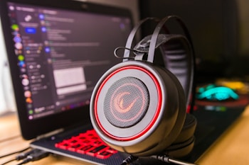 A pair of large over-ear headphones with a red and black design, positioned prominently in the foreground. In the background, there is a laptop with a visible chat application open on the screen, showing various text and icons. The laptop keyboard is illuminated with red backlighting, adding to the vibrant color scheme.