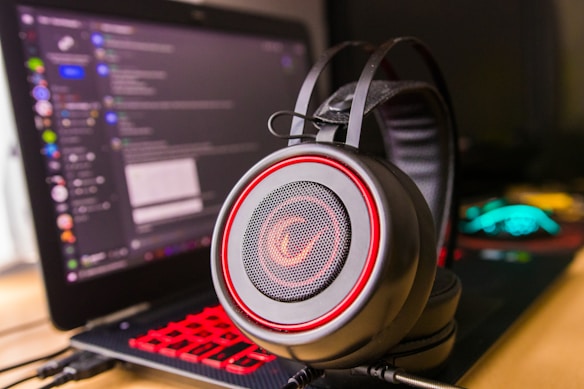 A pair of large over-ear headphones with a red and black design, positioned prominently in the foreground. In the background, there is a laptop with a visible chat application open on the screen, showing various text and icons. The laptop keyboard is illuminated with red backlighting, adding to the vibrant color scheme.