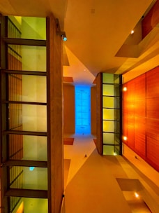 A symmetrical interior view featuring modern architectural elements with a strong emphasis on geometric shapes. The ceiling and walls are illuminated by warm, ambient lighting, casting an inviting glow. Glass partitions with metal frames create a contrast with wooden panels that add a rich texture. A central pane of blue-tinted glass draws the eye upwards, providing a cool contrast.