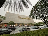 A modern cinema building features the 'Cinema XXI' sign in large gold letters. The front area is lined with palm trees and leafy greenery. Several cars are parked in the foreground, suggesting a parking area for visitors. The overcast sky suggests it might be early evening or a cloudy day.