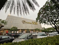 A modern cinema building features the 'Cinema XXI' sign in large gold letters. The front area is lined with palm trees and leafy greenery. Several cars are parked in the foreground, suggesting a parking area for visitors. The overcast sky suggests it might be early evening or a cloudy day.