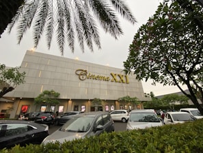 A modern cinema building features the 'Cinema XXI' sign in large gold letters. The front area is lined with palm trees and leafy greenery. Several cars are parked in the foreground, suggesting a parking area for visitors. The overcast sky suggests it might be early evening or a cloudy day.