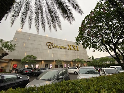 A modern cinema building features the 'Cinema XXI' sign in large gold letters. The front area is lined with palm trees and leafy greenery. Several cars are parked in the foreground, suggesting a parking area for visitors. The overcast sky suggests it might be early evening or a cloudy day.