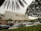 A modern cinema building features the 'Cinema XXI' sign in large gold letters. The front area is lined with palm trees and leafy greenery. Several cars are parked in the foreground, suggesting a parking area for visitors. The overcast sky suggests it might be early evening or a cloudy day.