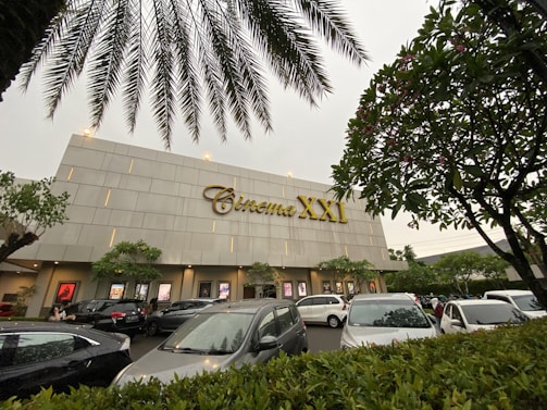 A modern cinema building features the 'Cinema XXI' sign in large gold letters. The front area is lined with palm trees and leafy greenery. Several cars are parked in the foreground, suggesting a parking area for visitors. The overcast sky suggests it might be early evening or a cloudy day.