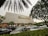 A modern cinema building features the 'Cinema XXI' sign in large gold letters. The front area is lined with palm trees and leafy greenery. Several cars are parked in the foreground, suggesting a parking area for visitors. The overcast sky suggests it might be early evening or a cloudy day.