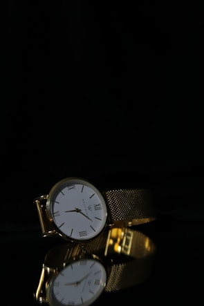 A luxurious gold wristwatch with a white face and Roman numerals is positioned on a reflective black surface. The watch features an intricate gold mesh strap and the reflection of the watch is clearly visible below it.