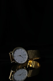 A luxurious gold wristwatch with a white face and Roman numerals is positioned on a reflective black surface. The watch features an intricate gold mesh strap and the reflection of the watch is clearly visible below it.