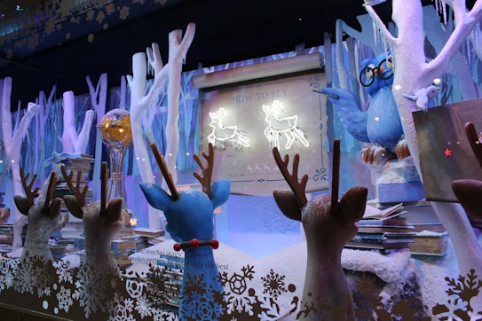 A whimsical winter scene featuring several reindeer figures facing a large owl figure wearing glasses. The setting includes snow-covered trees and stacks of books. A glowing sign reads 'How to Fly' above illustrated deer. The overall atmosphere is festive and magical.