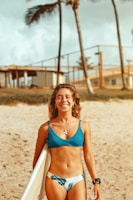 A woman with curly hair smiles while holding a surfboard on a beach. She is wearing a blue bikini with a palm tree pattern. In the background, there are palm trees, a fence, and beach huts, suggesting a tropical setting.