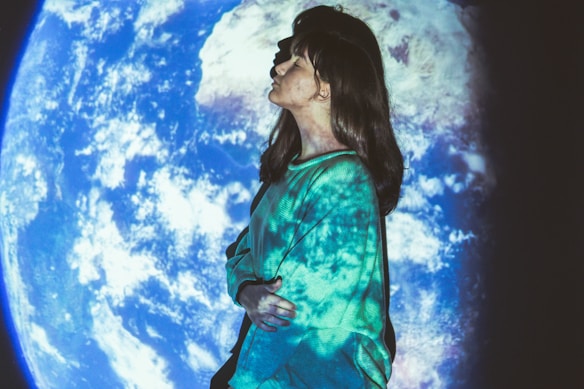 A person is in profile, standing against a backdrop of Earth projected onto a screen. The person is wearing a teal sweater, and their face and clothing are also covered in the Earth projection, creating an ethereal look.
