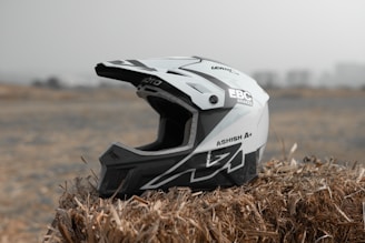 A black and white motocross helmet is resting on a bale of hay in an outdoor setting with a blurred background. The helmet has various brand logos and text decals, featuring sharp geometric designs.